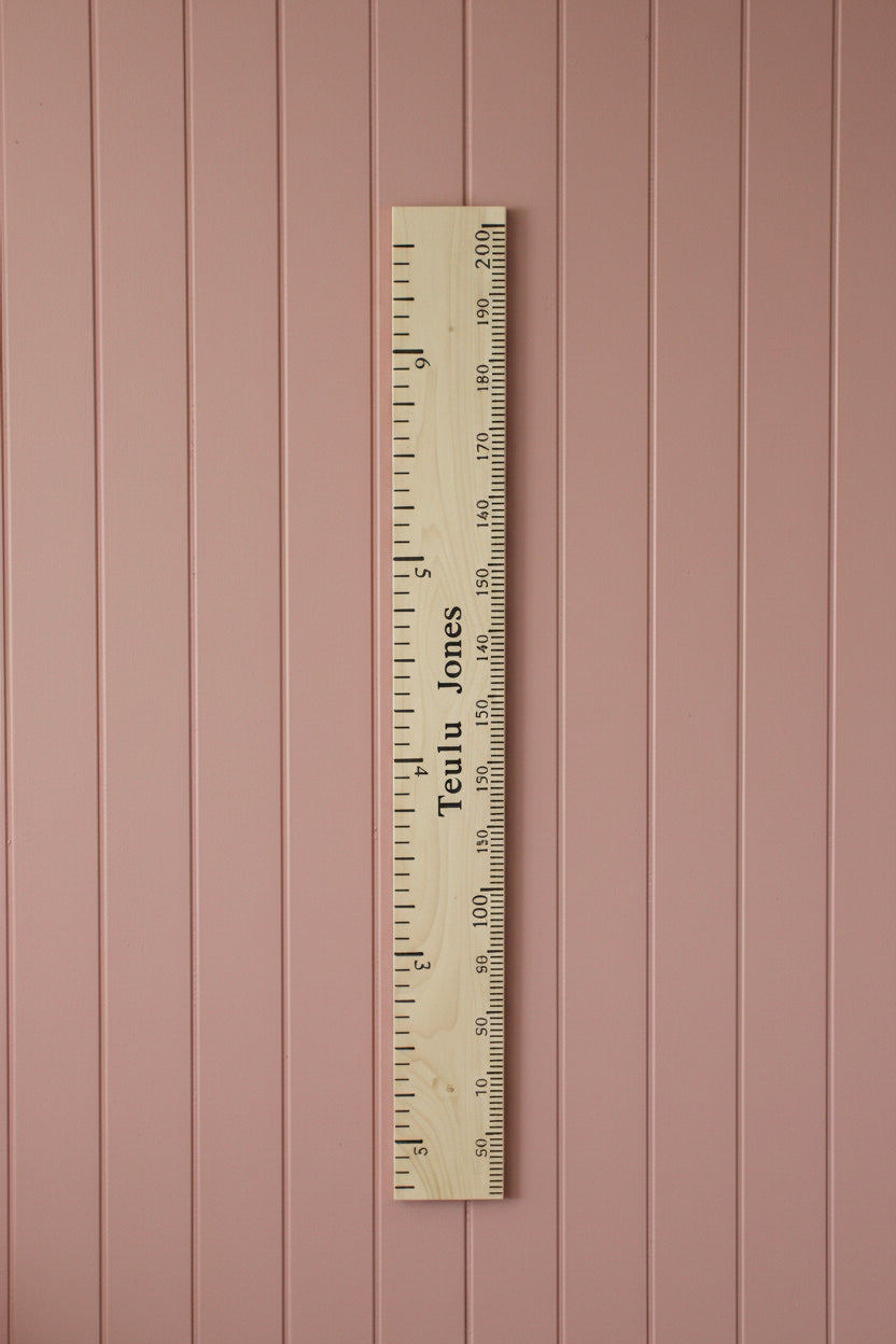 A tall wooden wall ruler with black printed measurements and the text 'Tulips Jones' down the center, leaning against a white wall in a cluttered workshop or storage room.