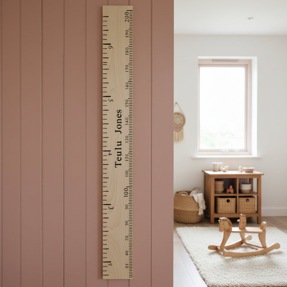 A personalized wooden floor height marker with the name 'Teelu Jones' printed vertically, shown standing on a wooden floor against a white wall in a workshop setting.