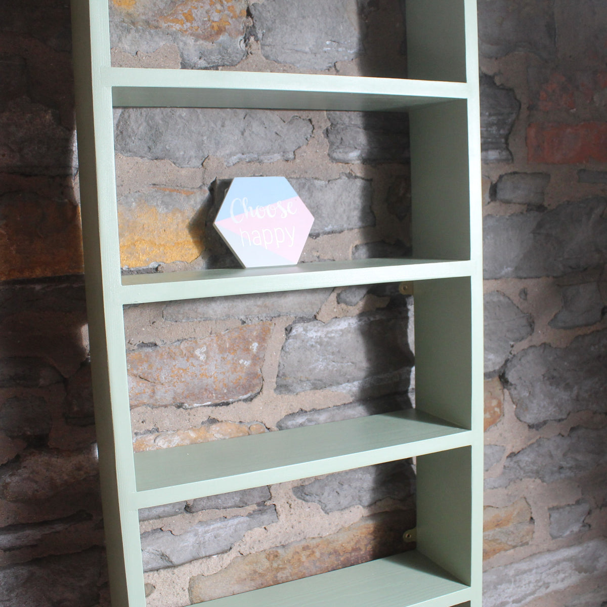 A tall, narrow sage green shelving unit with four open shelves stands against a rustic stone wall. A small hexagonal sign reading 'choose me again' rests on the second shelf.
