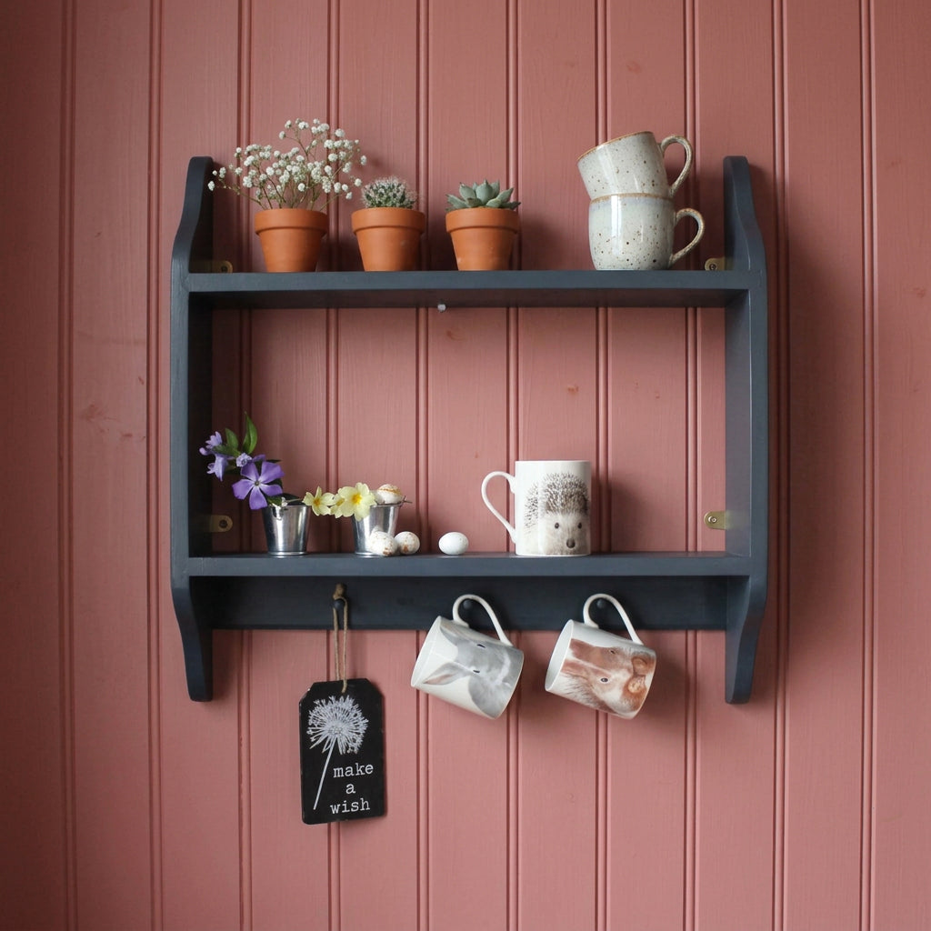 A dark gray wooden open shelf mounted on a light gray wall, styled with small metal pots, flowers, a mug, two hanging mugs, and a small black sign.