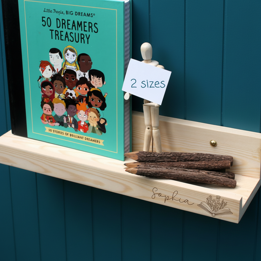 A wooden floating shelf mounted on a teal paneled wall, styled with a turquoise book titled "50 Dreamers Treasury," a small wooden mannequin holding a "2 sizes" sign, three wooden pencils, and a custom engraved nameplate reading "Sophia.".