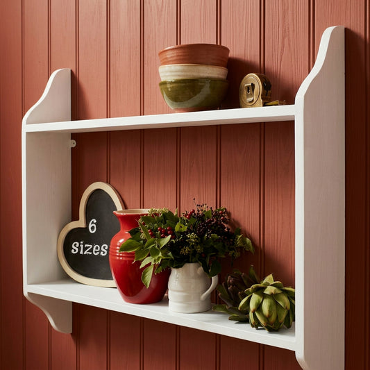 A light gray painted wooden open shelf mounted on a white wall, styled with a red vase, a white pitcher filled with greenery, a heart-shaped chalkboard sign reading '6 sizes', and a green artichoke.