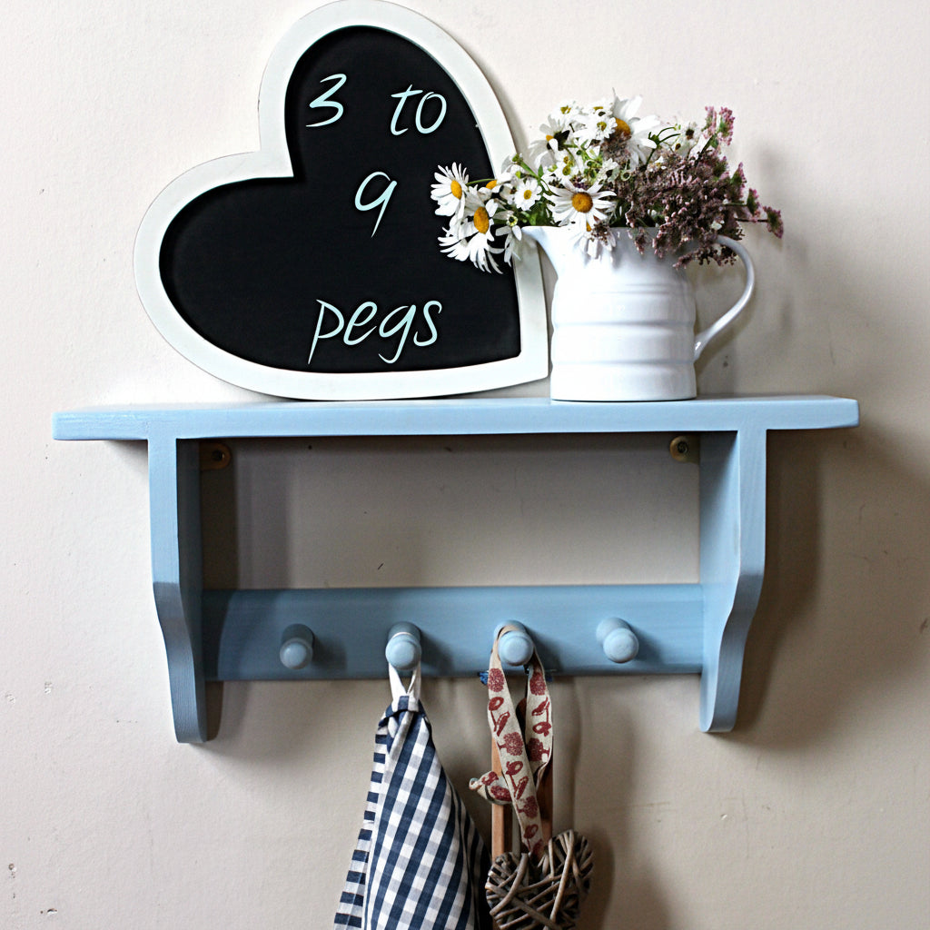 A light blue wooden wall-mounted shelf with two hooks and a heart-shaped chalkboard on top, styled with a white pitcher of flowers against a white hexagonal tile wall.