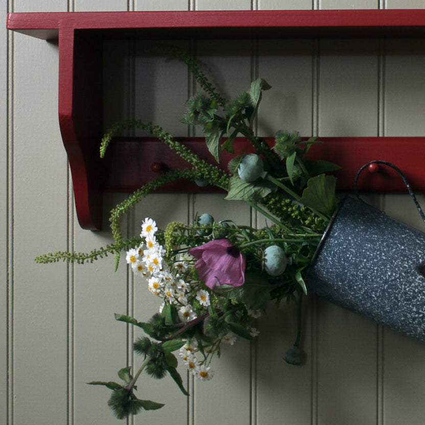 A rustic decorative arrangement featuring a blue speckled metal bucket hanging from a red wooden shelf, filled with artificial green foliage, white daisies, and a pink flower, set against a plain off-white wall.