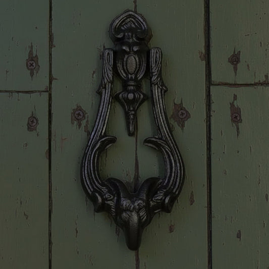 An ornate black metal door knocker featuring a ram's head at the bottom and a decorative top section, mounted on a dark green paneled door.