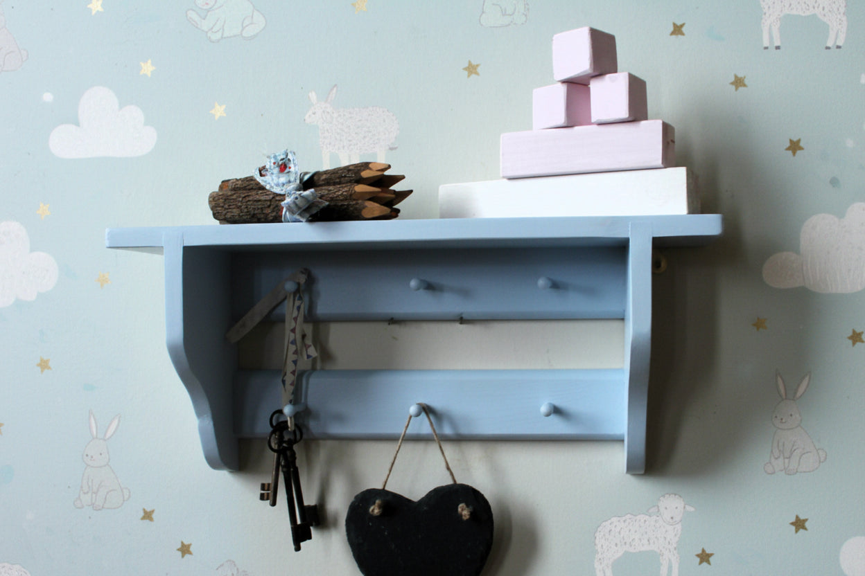 A light blue wooden wall shelf mounted on a beige wall, styled with stacked pink and white blocks, a small decorative object, keys hanging from hooks, and a black heart-shaped ornament.