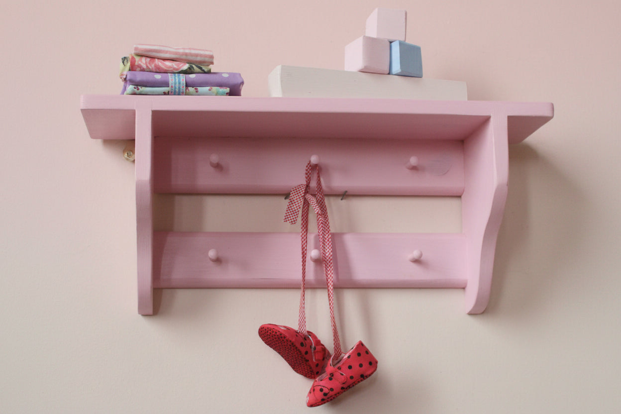 A pink wooden wall-mounted shelf with two small pegs and a hanging red-and-white polka dot ribbon, with red-and-white polka dot shoes dangling below. Books and stacked blocks sit on top against a plain light wall.