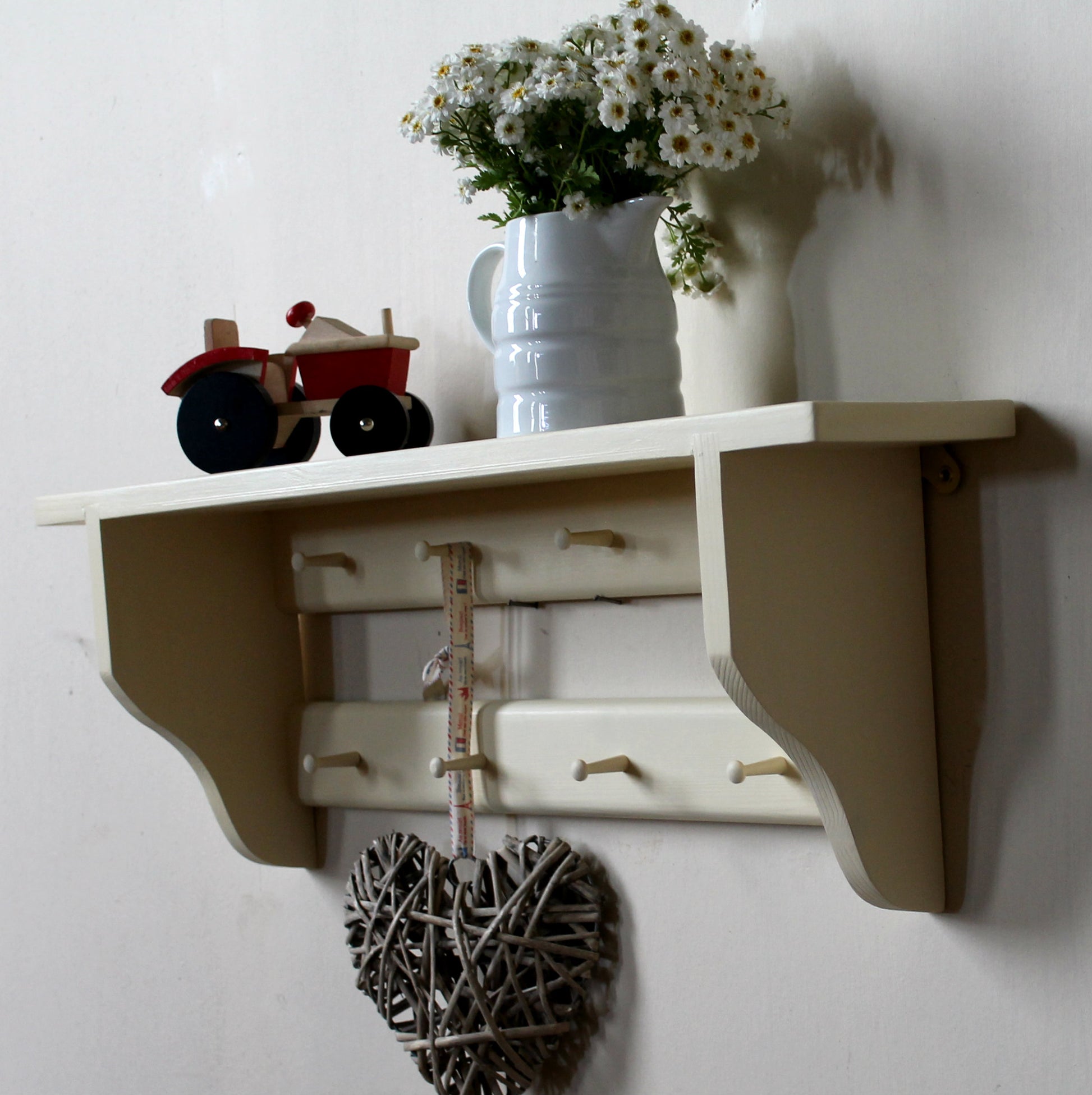 A cream-colored wall-mounted wooden shelf with hooks and a hanging twig heart ornament, styled with a white pitcher of daisies and a small red toy tractor against a white wall.