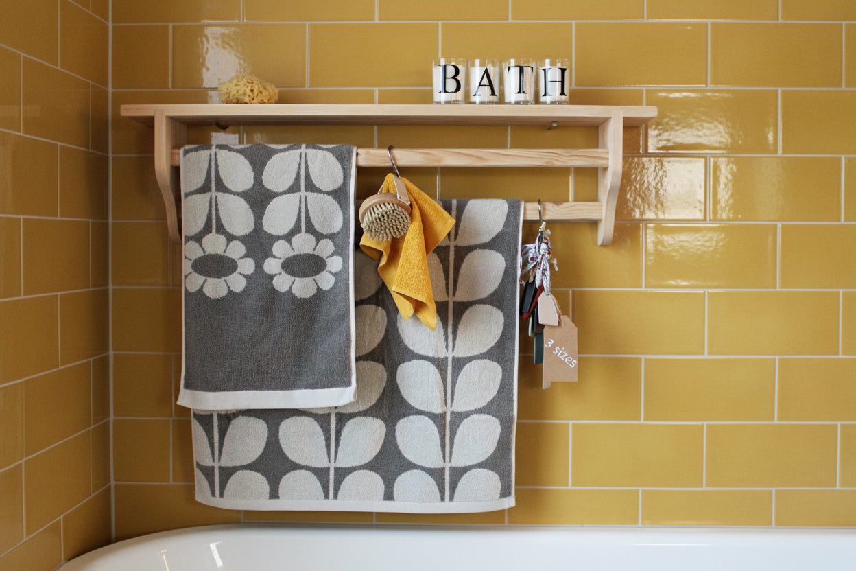 A light natural wood wall-mounted shelf with a horizontal bar, shown holding three patterned bath towels, a yellow hand towel, a sponge, and small glass cups on a shelf. The wall is a neutral light gray.