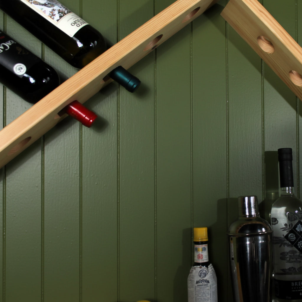 A light natural wood wall-mounted wine rack with circular cutouts, shown holding several wine bottles against a green wall. Additional bottles and bar accessories are partially visible in the lower right corner.