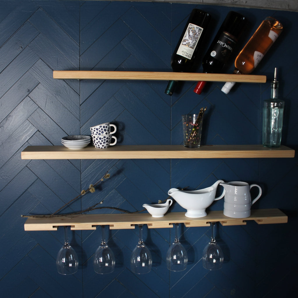 Three light natural wood floating shelves mounted on a blue wall, styled with wine bottles, dishes, cups, and hanging wine glasses.