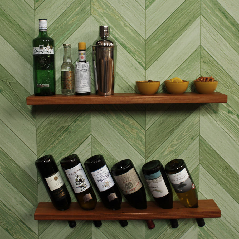 Two wooden floating shelves mounted on a white wall, styled with liquor bottles, a cocktail shaker, small bowls, and wine bottles resting horizontally in the shelf holders.