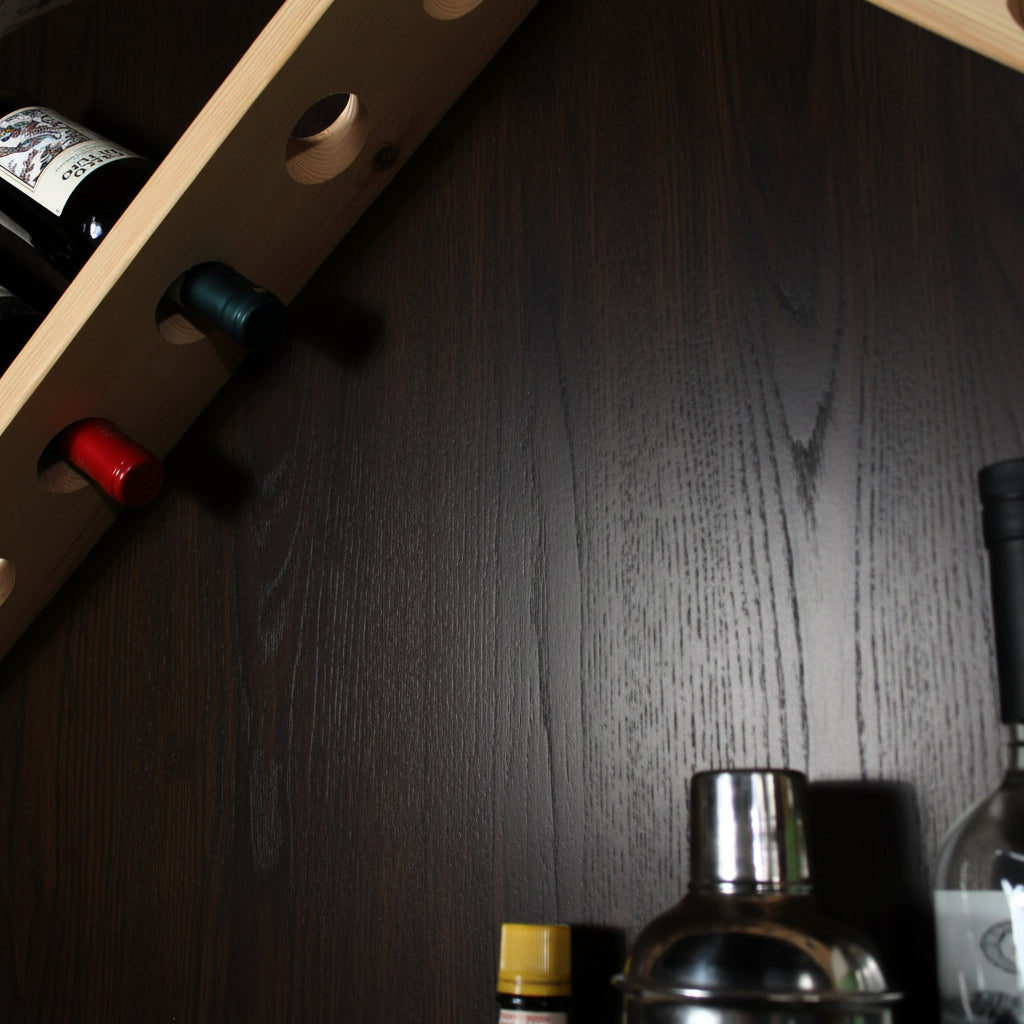 A close-up of a light natural wood wine rack mounted on a dark wall, holding several wine bottles. The photo is cropped tightly, with the rack entering from the top-left corner and cocktail shaker and other bar items partially visible at the bottom-right.