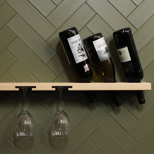 A wall-mounted wooden wine rack home bar shelf with concealed brackets, shown holding three wine bottles on top and three clear wine glasses hanging from the side, against a green wall. 