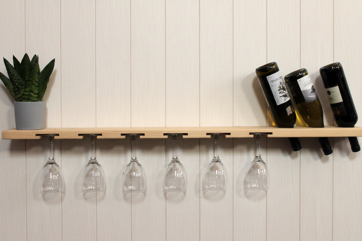 A light wood wall-mounted shelf with integrated wine glass hangers and bottle holders, shown against a white wall with six inverted wine glasses hanging below and three wine bottles resting on the right side. A small potted succulent sits on the left.
