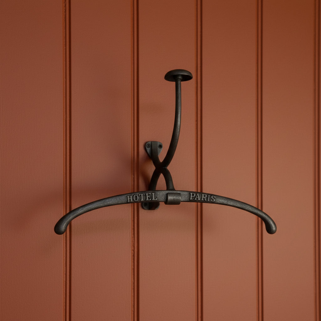 A dark metal wall hook with a curved bar engraved with 'HOTEL PARIS', displayed against a vintage black-and-white toile wallpaper pattern.