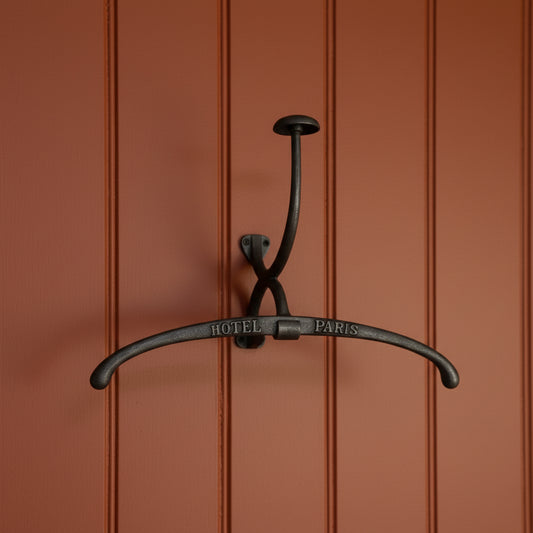 A dark metal wall hook with a curved bar engraved with 'HOTEL PARIS', displayed against a vintage black-and-white toile wallpaper pattern.