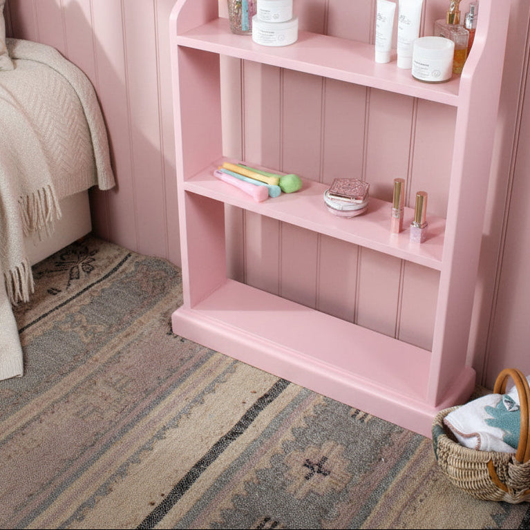 A light pink three-tier wooden shelf with decorative items on its shelves, set against a gray brick wall on a patterned rug with a woven basket nearby.