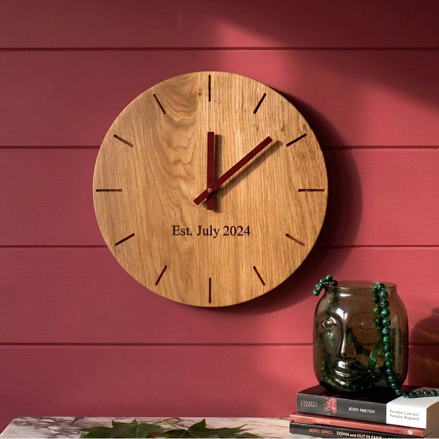 A round wooden clock with dark red hands and engraved text reading 'Est. July 2024' sits on a light wood block with green leaves on top.