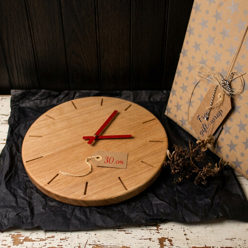 A round wooden wall clock with red hands and minimalist cutout hour markers, displayed on black tissue paper next to a wrapped gift box and dried flowers. A small tag on the clock reads 30 cm.