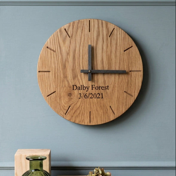 A round wooden wall clock with a natural oak grain finish, custom engraved text reading 'Dalby Forest 3/6/2021', and dark metal hands, styled on a white background with decorative blocks and a green glass bottle.