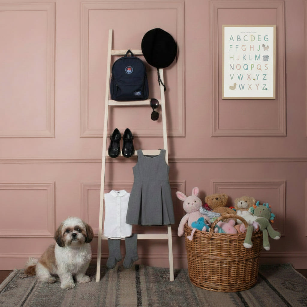 A small white and brown dog sits on a patterned rug next to a light wood ladder shelf styled with a school black hat, a black school bag, black school shoes, a school uniform, against a pink wall panel. 