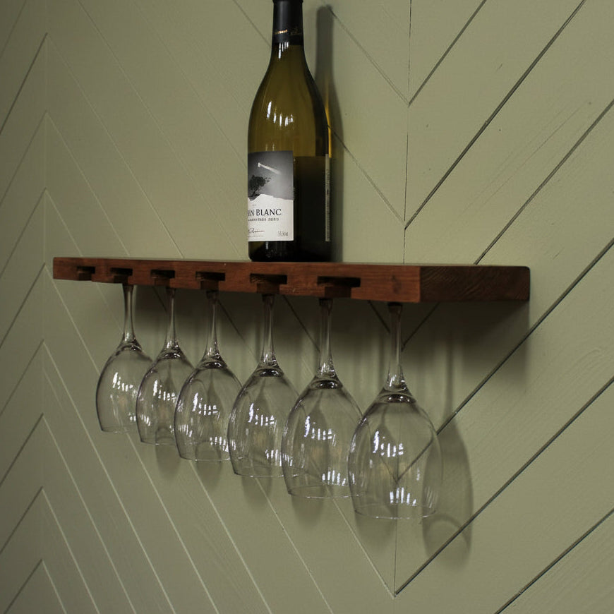 A dark brown wooden wall-mounted shelf with five hanging wine glass holders, shown against a white wall with a wine bottle and five clear wine glasses displayed on and below the shelf.