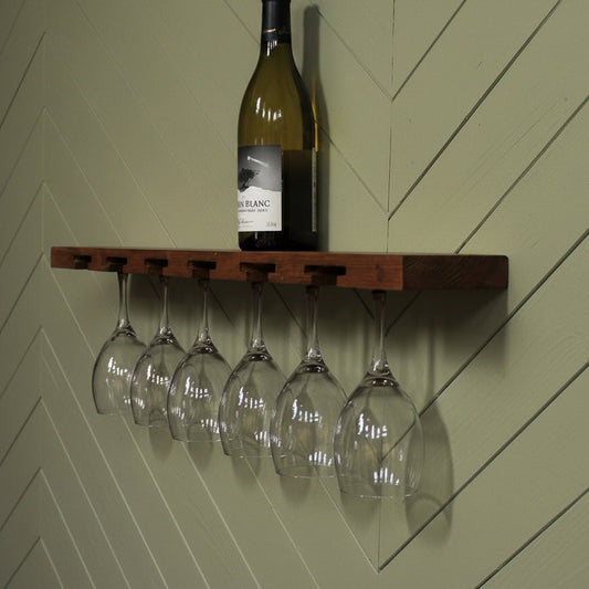 A dark brown wooden wall-mounted shelf with five hanging wine glass holders, shown against a green wall with a wine bottle and five clear wine glasses displayed on and below the shelf.