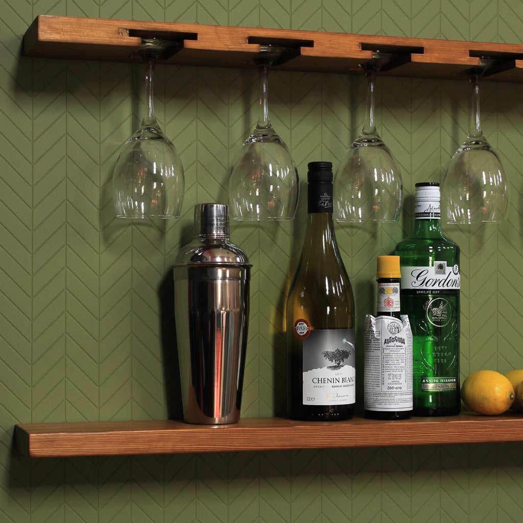 A wooden wall-mounted bar shelf with a matching top rack holding five wine glasses, styled with a cocktail shaker, wine bottles, and lemons against a white wall.