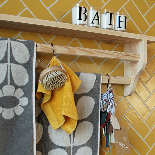 Bathroom shelf with towel rail holding a patterned towel and wooden brush, shelf above decorated with pink soap and glass candle holders spelling 'BATH'