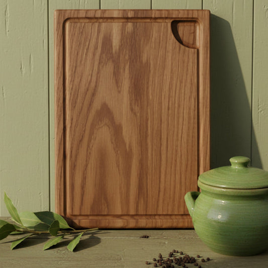 A rectangular wooden cutting board with rounded corners and a curved handle cutout in the top right, standing upright on a rustic wooden surface next to a green ceramic lidded pot and a sprig of green leaves.